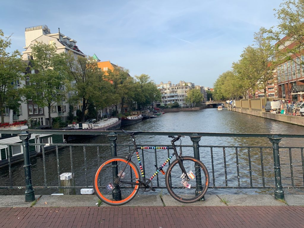 A single speed bike with a rainbow-striped black frame in front of a canal.